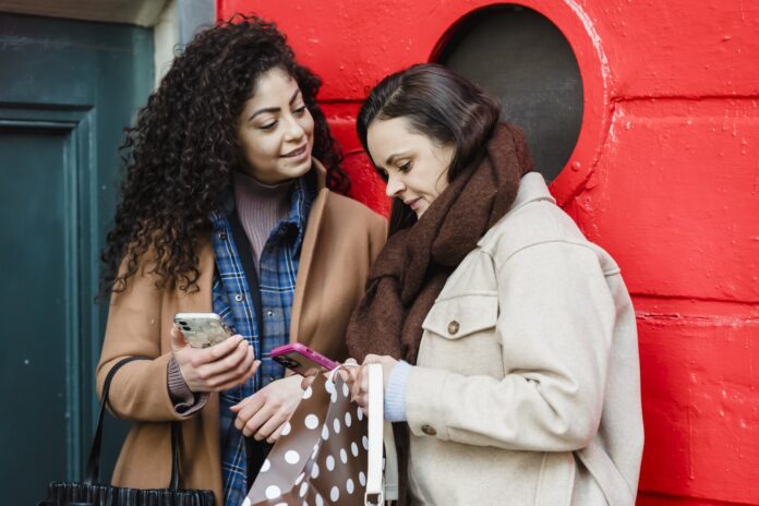 Positive young diverse female best friends in stylish warm clothes standing on city street near red door and browsing mobile phones
