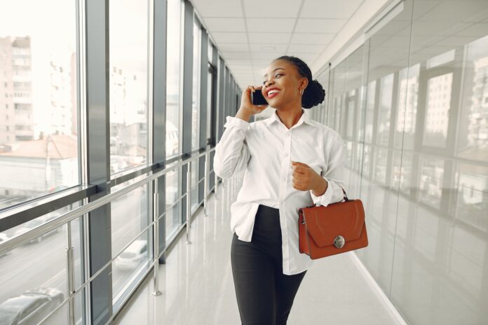 Happy black stylish female office worker in formal wear having conversation on mobile phone while strolling along modern building corridor and looking out window