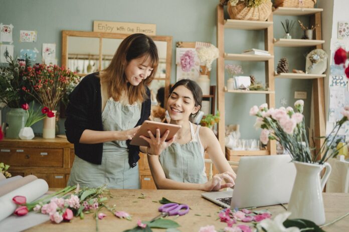 Content multiethnic female florists in aprons at table with laptop and browsing tablet while preparing for order in creative floristry studio