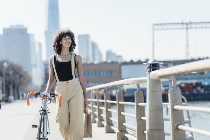 Young ethnic female in casual clothes walking with bike on city seafront and smiling while looking away