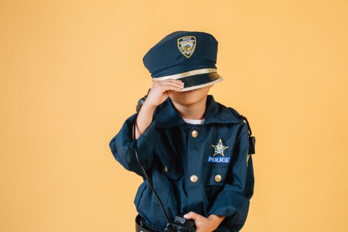 Unrecognizable child in police uniform standing in studio with transceiver in hand and pulling cap over face on yellow background